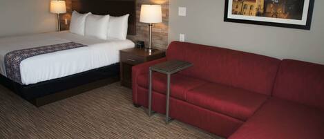 A hotel room with a bed, a red sofa, a nightstand with a lamp, and a framed picture on the wall.