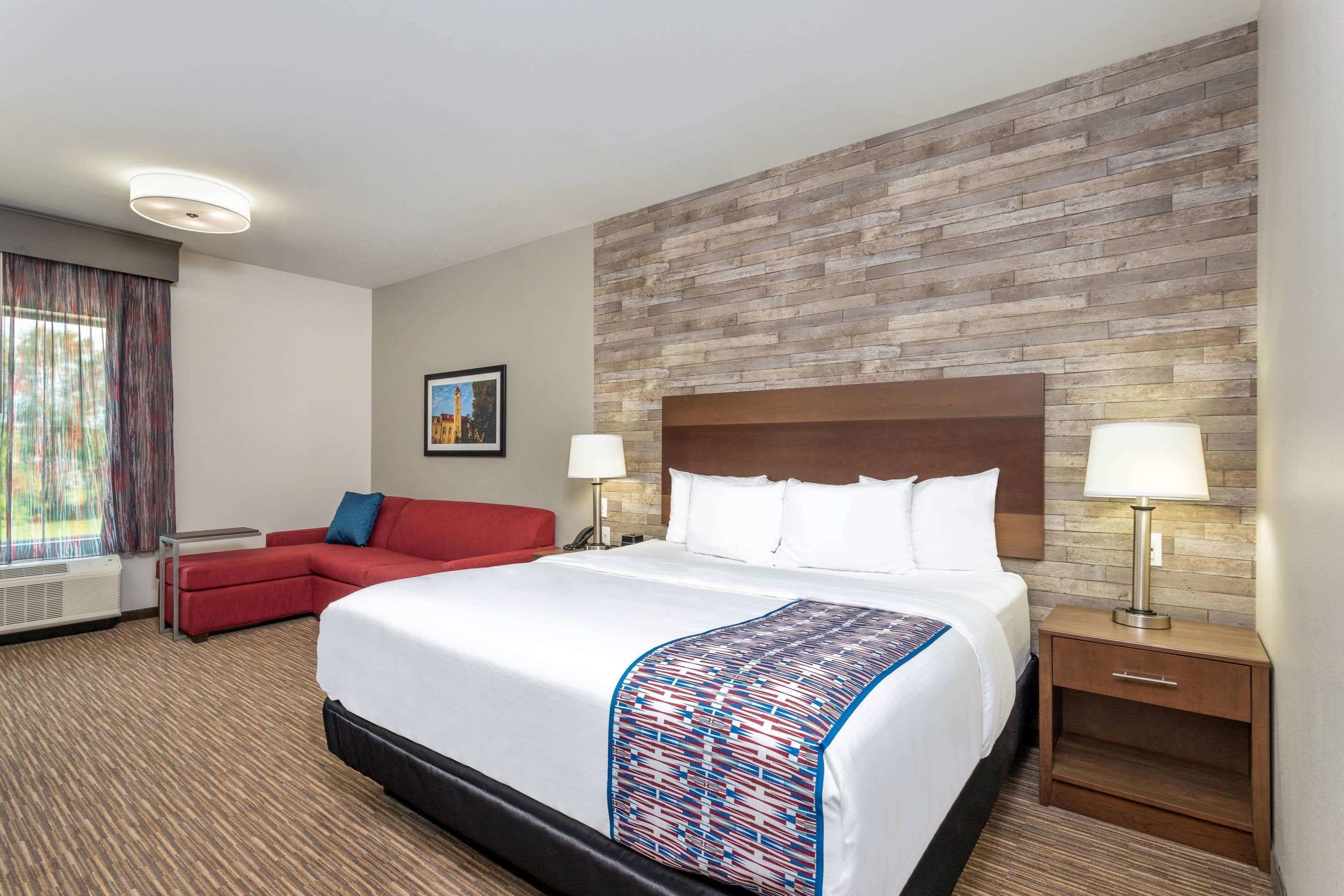 A hotel room with a large bed, a red sofa, a nightstand with a lamp, and a window with curtains.