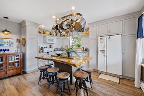 Eine Küche mit weißen Schränken, einer zentralen Kochinsel mit Barhocker, einer Pendelhängung für Kochtöpfe und einem Kühlschrank.