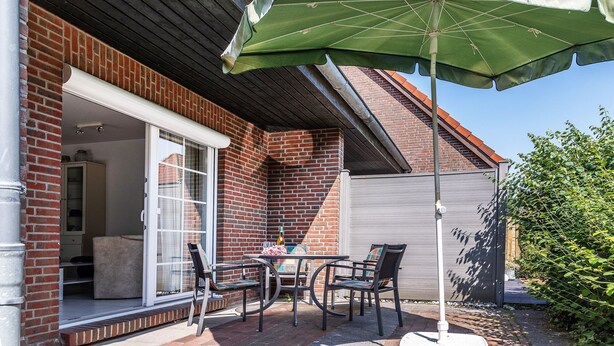 A patio with a table and chairs, a green umbrella, and a brick house with a garage door. - Timeless and elegant: the semi-detached house impresses with both its brightness (Carolinensiel)