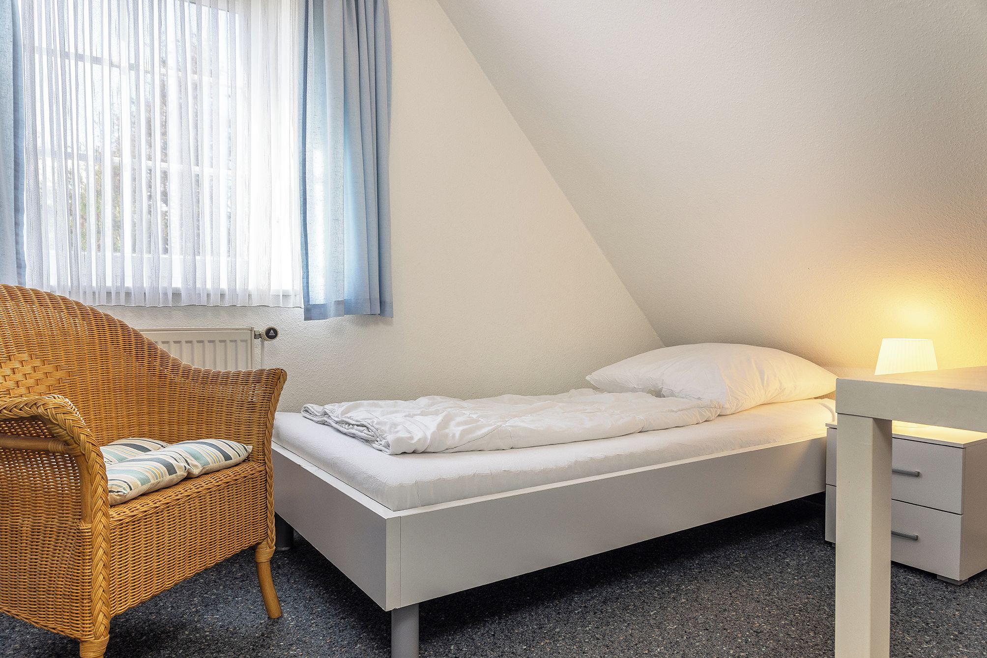 A small, single-bed room with a sloped ceiling, a wicker chair with cushions, a bedside table with a lamp, and a window with sheer curtains.