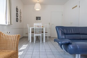 Un salon avec un canapé bleu, un fauteuil en osier, une table à manger avec des chaises blanches et un luminaire suspendu.