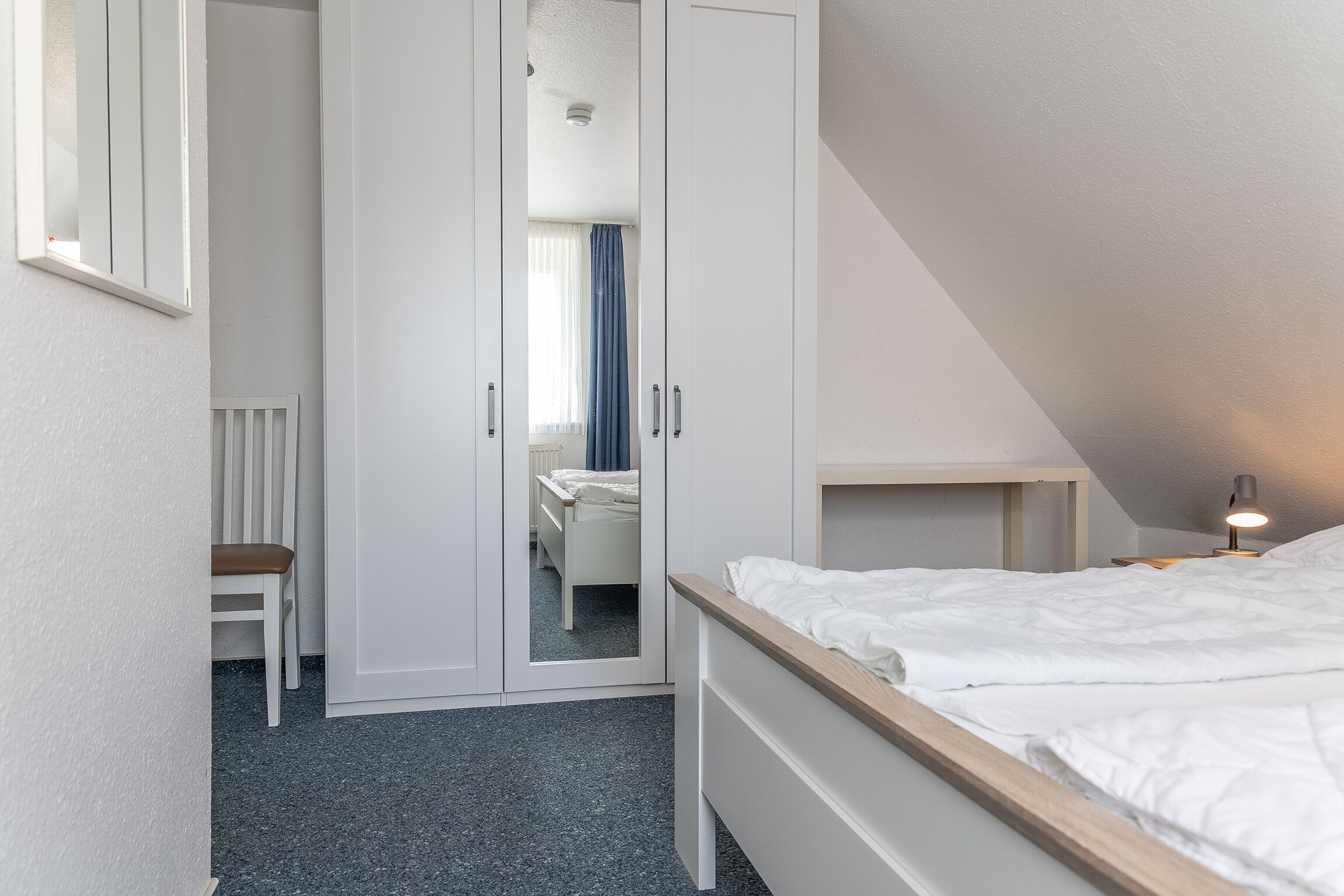 Et soveværelse med seng, garderobeskab, stol og skrivebord med lampe.