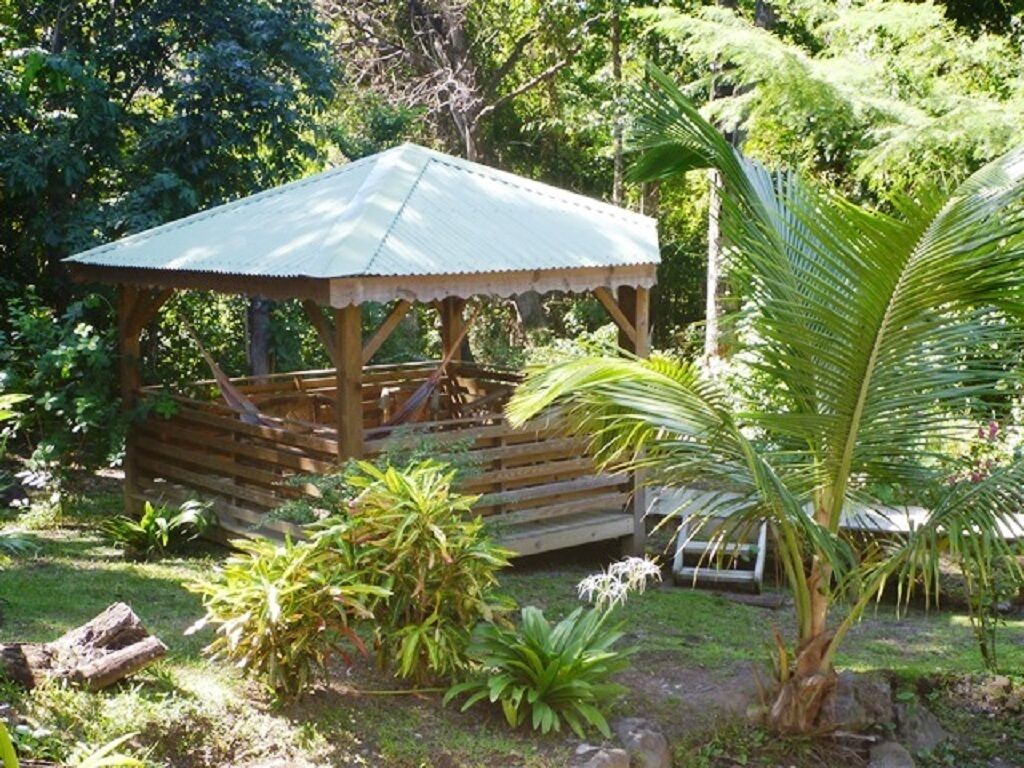 Ein hölzerner Pavillon mit Wellblechdach, umgeben von üppigem Grün und einer Palme.
