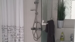 Casa de banho com chuveiro em cabine de vidro, lavatório branco, espelho e planta em vaso.