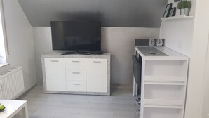 Um espaço compacto com armário branco, televisão de tela plana, mesa pequena com duas taças de vinho e um radiador.