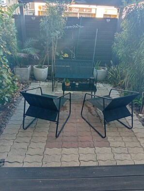 An outdoor seating area with two black metal chairs and a small table, surrounded by potted plants and a paved floor.
