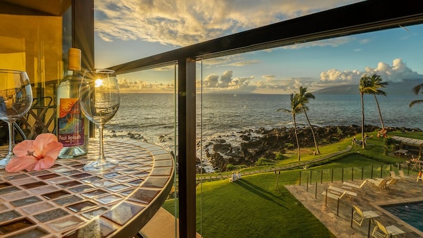 A balcony with a view of the ocean, a table with wine glasses and a bottle, and a tropical flower.