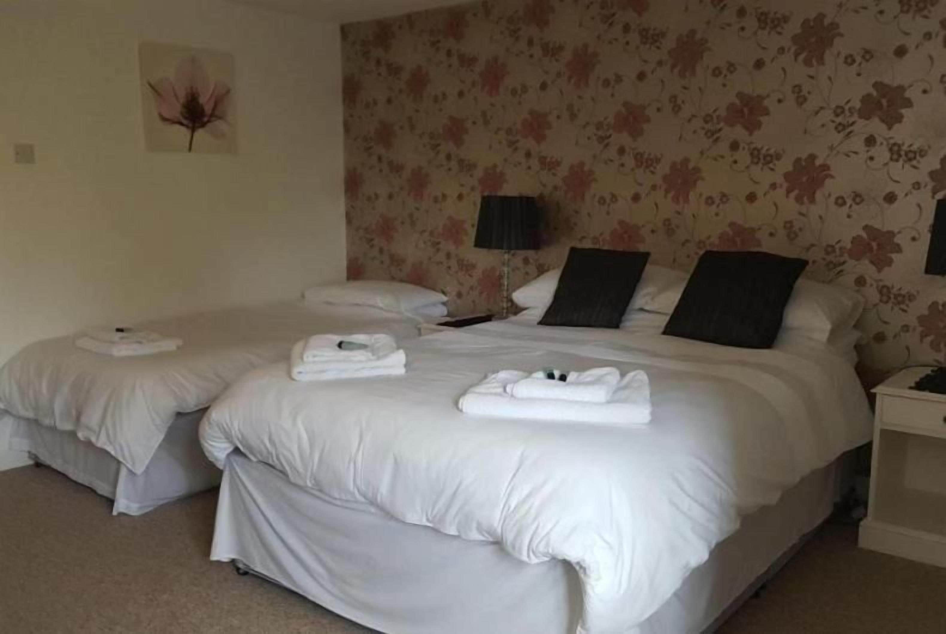 A hotel room with two beds, each with white linens and towels, a nightstand with a lamp, and a floral wallpaper pattern.