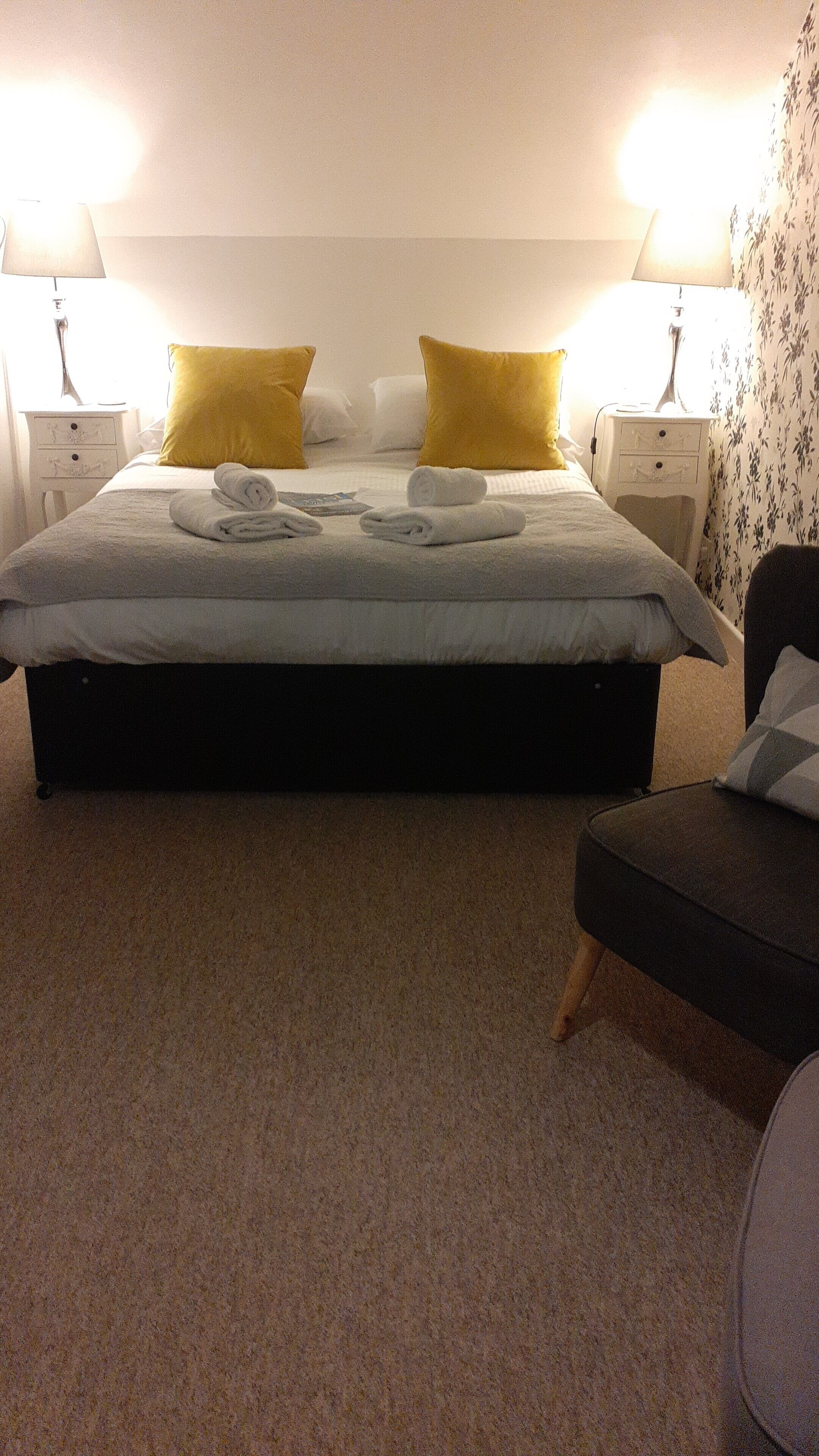 A neatly made bed with white linens and two yellow pillows, a small bedside table with a lamp, and a chair with a cushion.