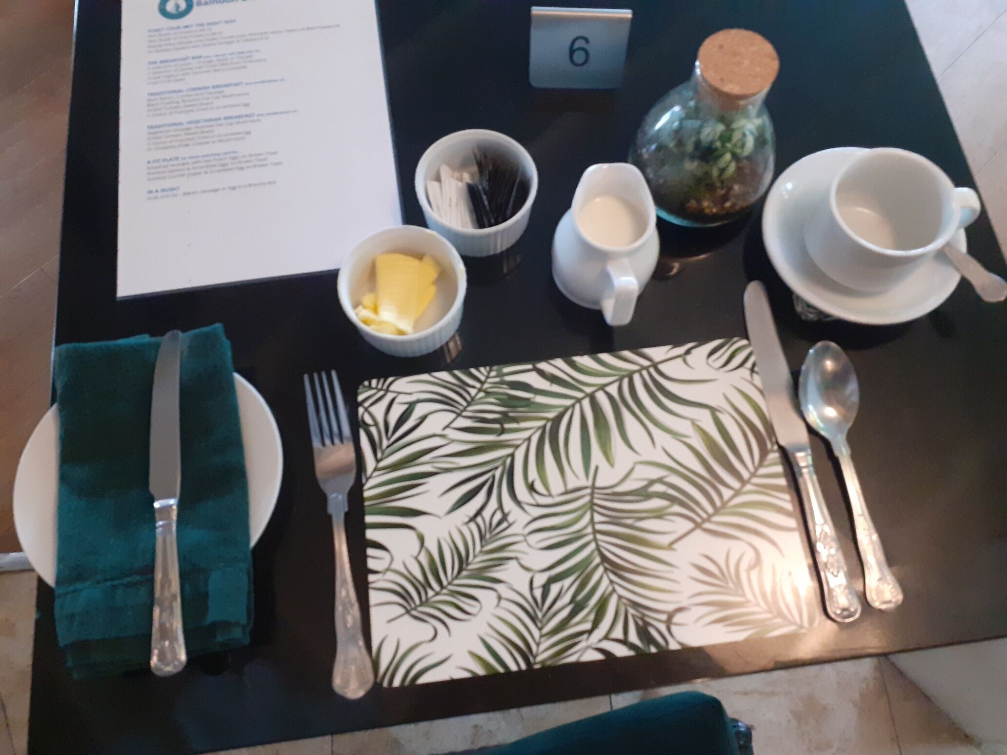 A table set with a green napkin, a fork, a knife, a spoon, a teapot, a cup, and a small bowl of butter.