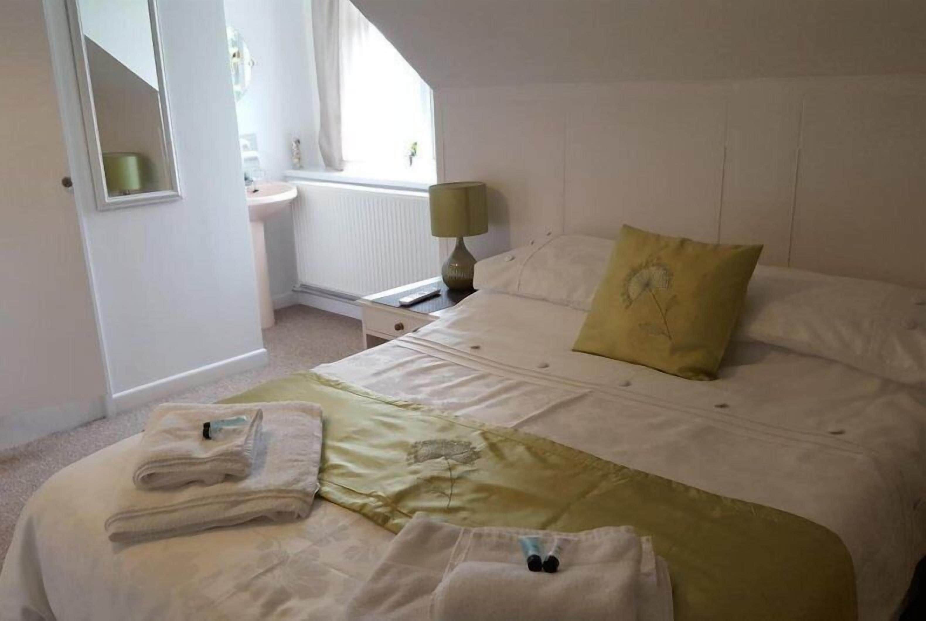A hotel room with a bed, a bedside table with a lamp, a window, and a bathroom visible in the background.