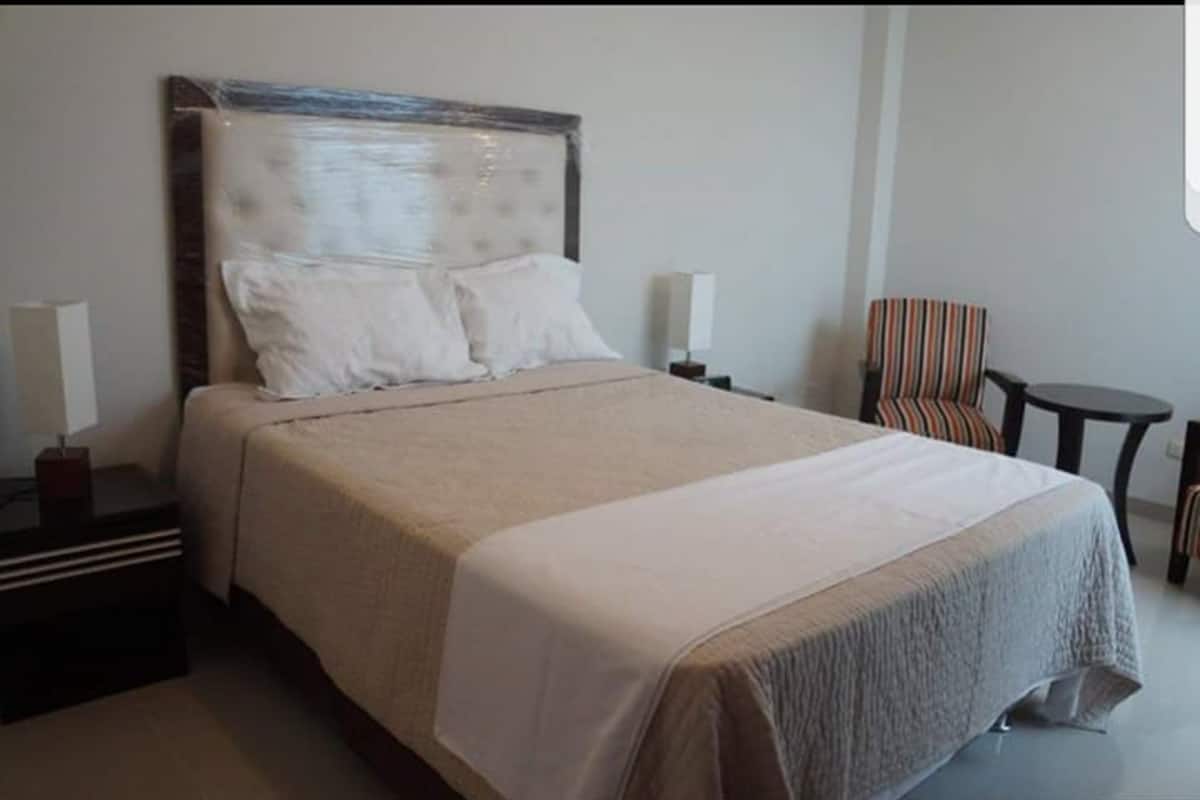 A neatly made bed with a headboard, two bedside tables with lamps, a striped armchair, and a small round table.