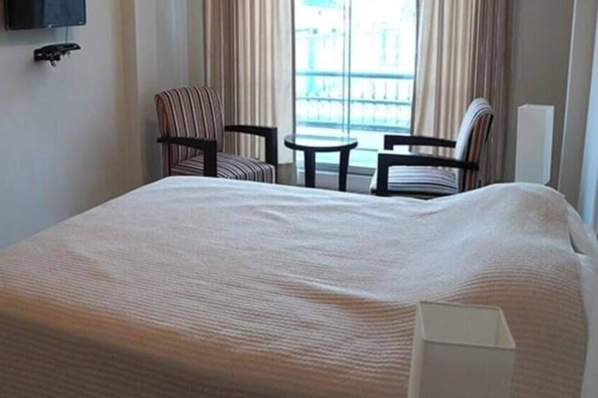 A hotel room with a large bed, two chairs, a small table, a TV mounted on the wall, and a window with curtains.