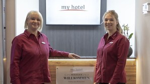 Two receptionists at a hotel reception desk with a sign that reads "my hotel" and "HERZLICH WELCOME".