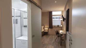 Quarto de hotel moderno com chuveiro, escrivaninha com cadeira, radiador e janela com cortinas.