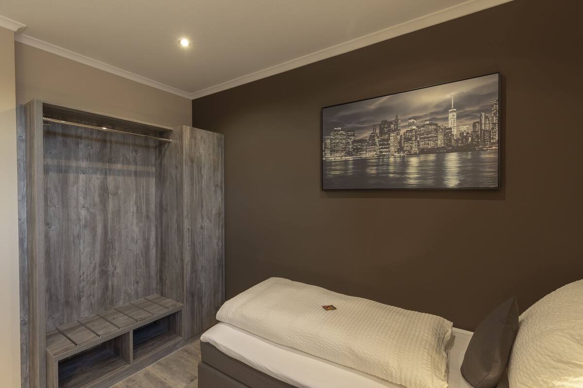 A bedroom with a bed, a bench, a wardrobe, and a cityscape painting on the wall.