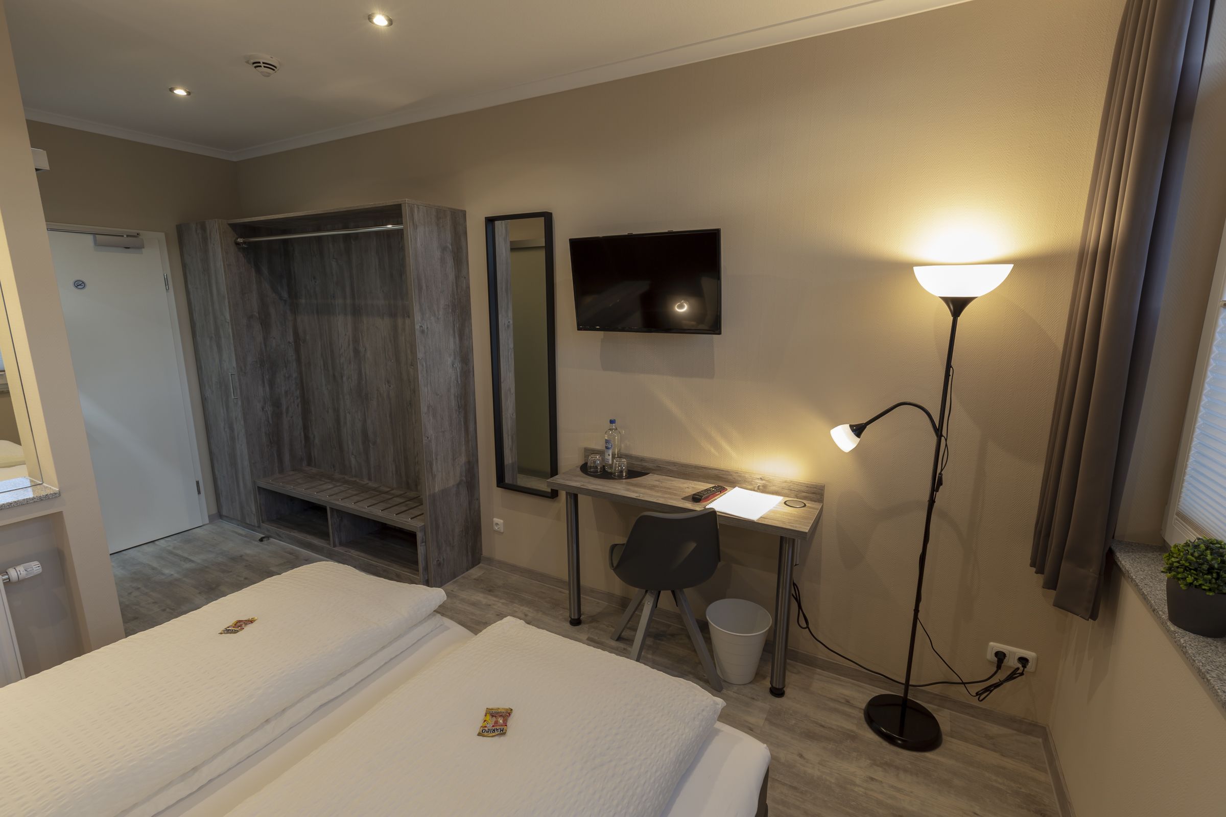 Ein Hotelzimmer mit zwei Betten, einem Schreibtisch mit Computer, einem Spiegel, einem Fernseher, einer Lampe und einer kleinen Pflanze.
