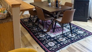 Uma sala de jantar com uma mesa de madeira, seis cadeiras, um tapete estampado e um quadro na parede.