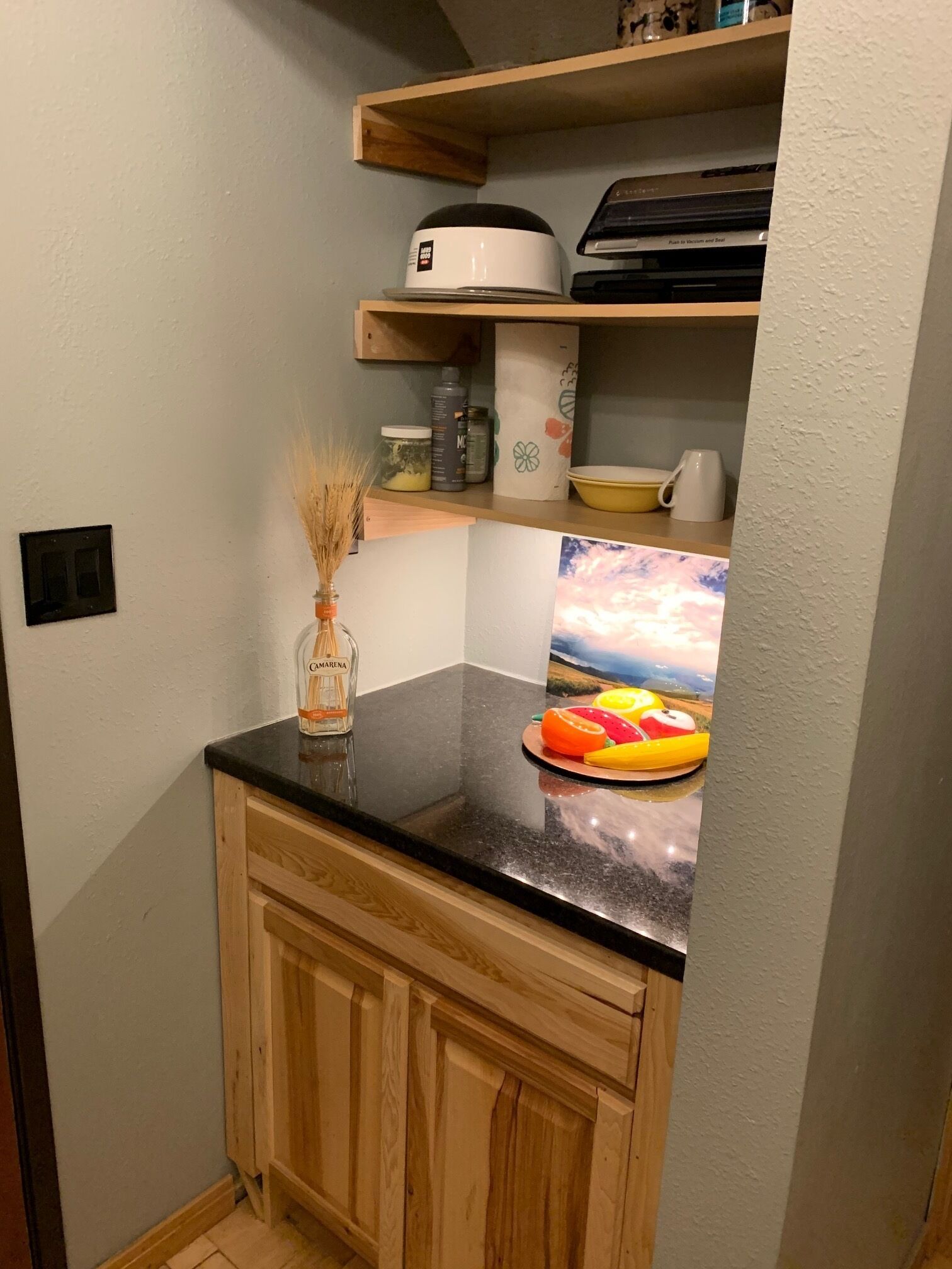 A kitchen with wooden cabinets, a black countertop, and a fruit bowl. There is a painting of a landscape on the wall.