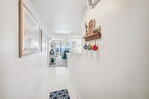 A hallway with a tiled floor, a white wall with a shelf holding decorative items, and a door leading to a balcony with a view.