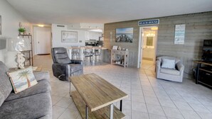 A living room with a sofa, a wooden coffee table, and a dining area with chairs and a table. There is a kitchen in the background and a sign that reads "BEACH HOUSE".