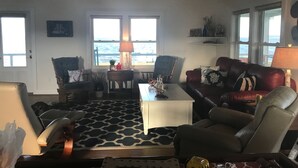 Una sala de estar con un tapete estampado, una mesa de centro, un sofá y dos sillones. Hay ventanas que dan al mar.