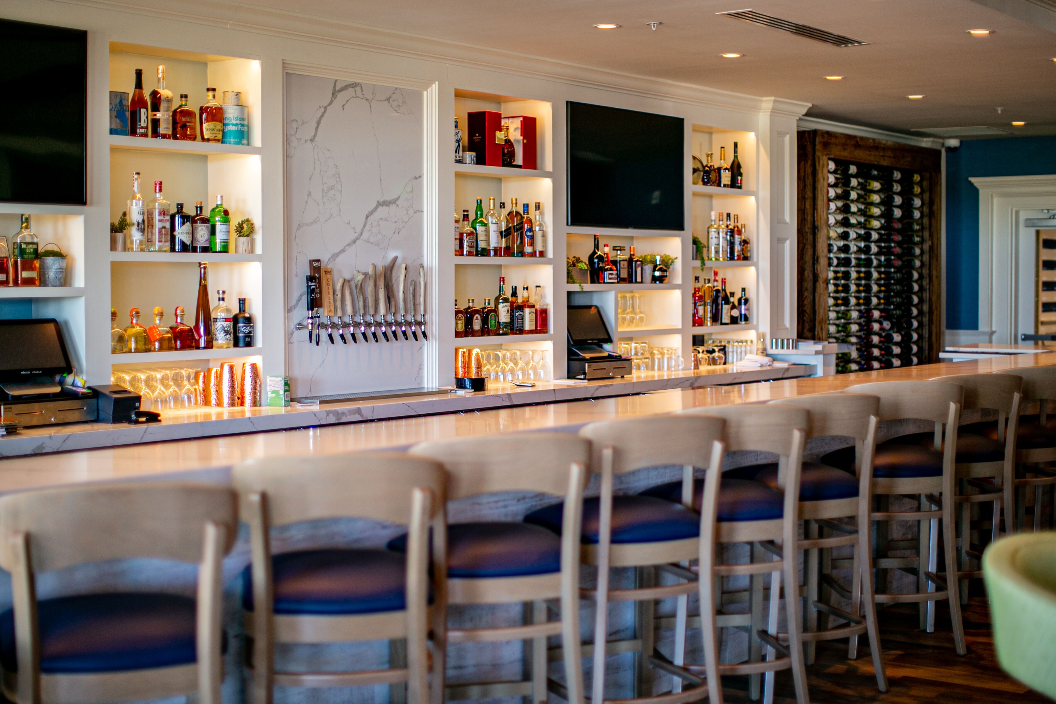 A bar area with multiple shelves of liquor bottles, a bar counter, and several bar stools.