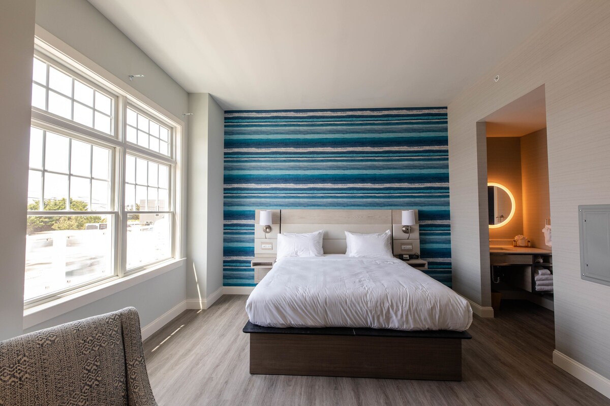Une chambre d’hôtel moderne avec un grand lit, une vue sur l’extérieur par une fenêtre, un mur à rayures bleues et une petite salle de bain.