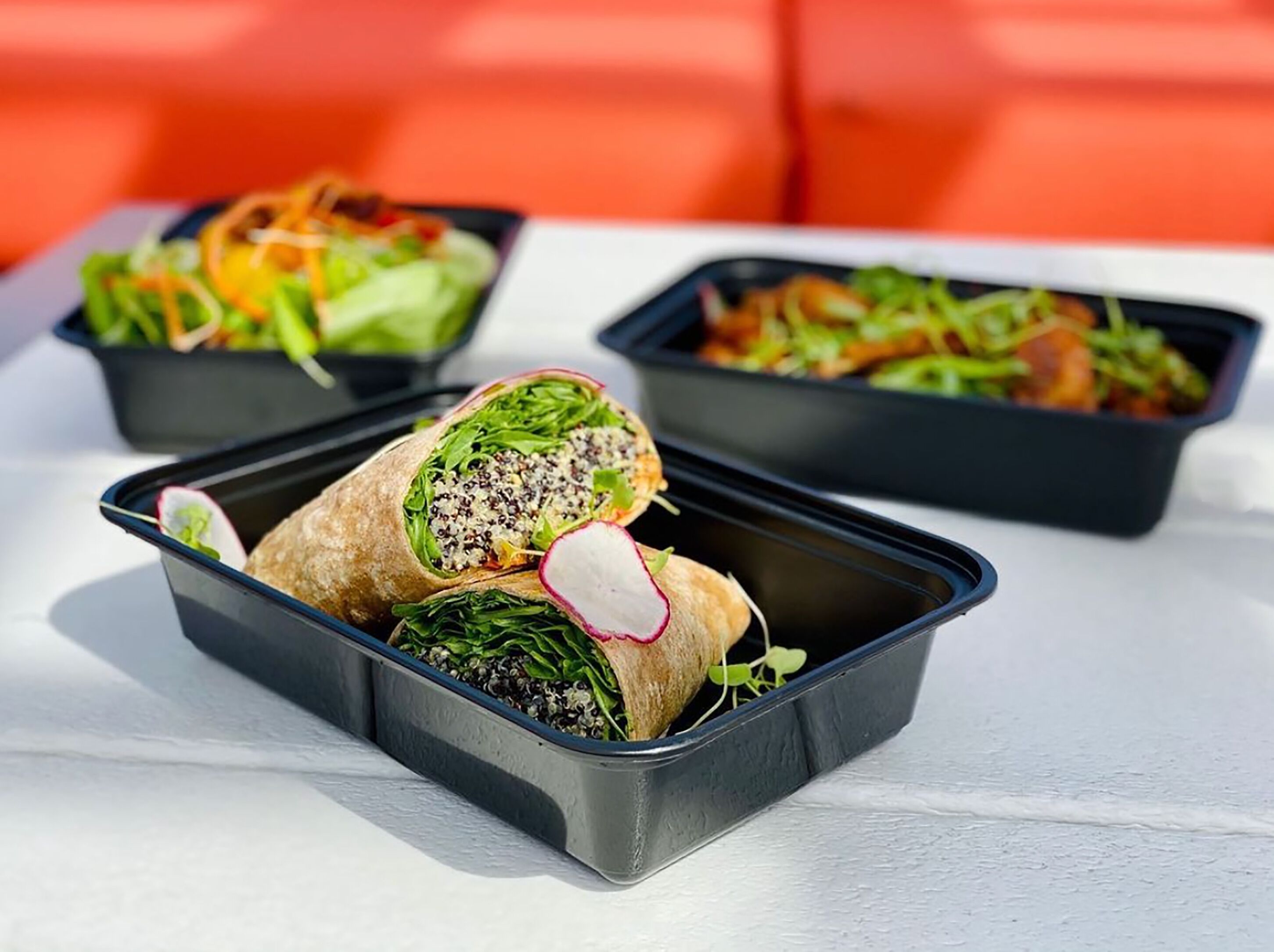 Drei schwarze, rechteckige Behälter mit Wraps und Salaten auf einer weißen Oberfläche.