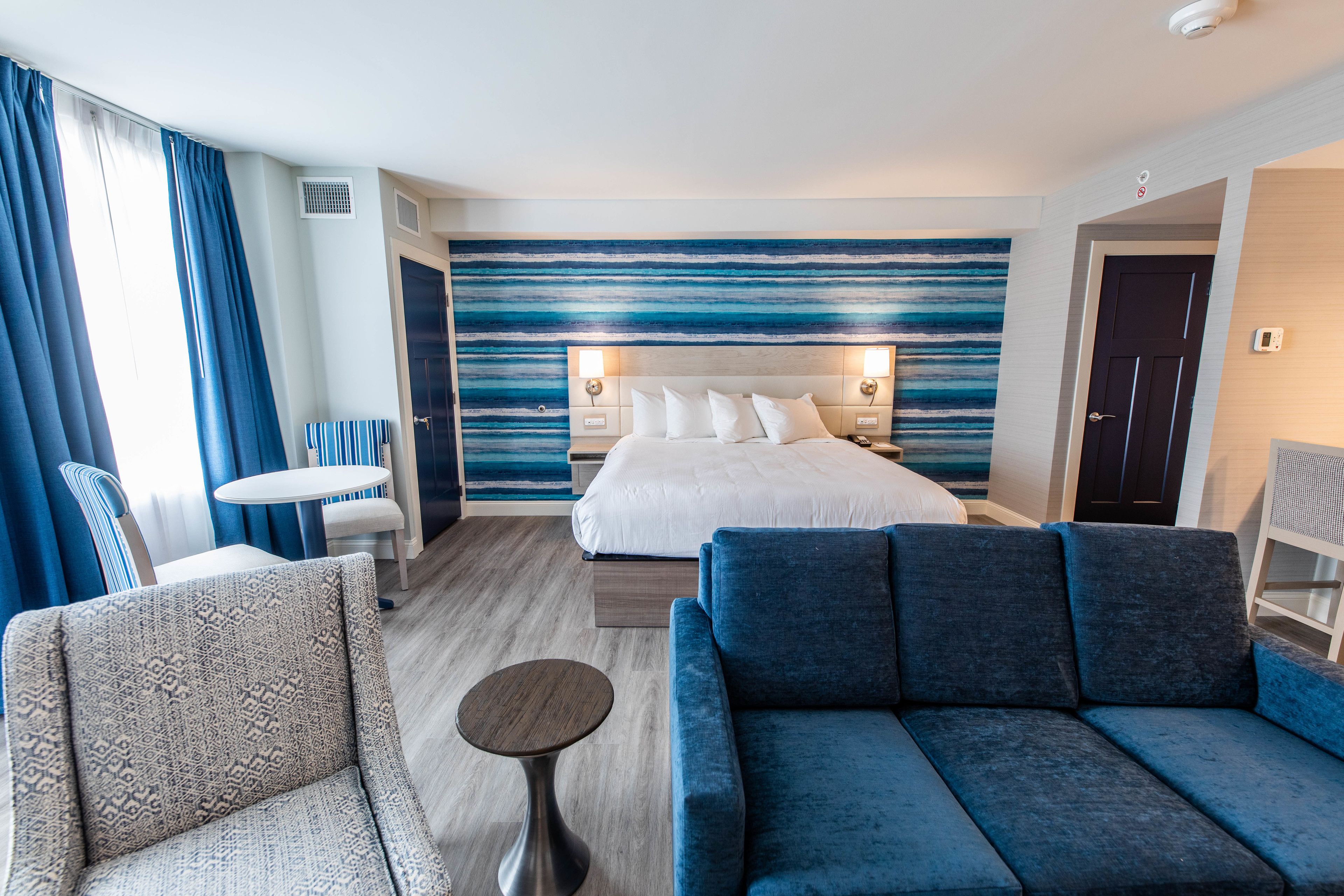 Et hotelværelse med en seng, en blå sofa, en mønstret lænestol, et lille rundt bord og havudsigt fra vinduet.