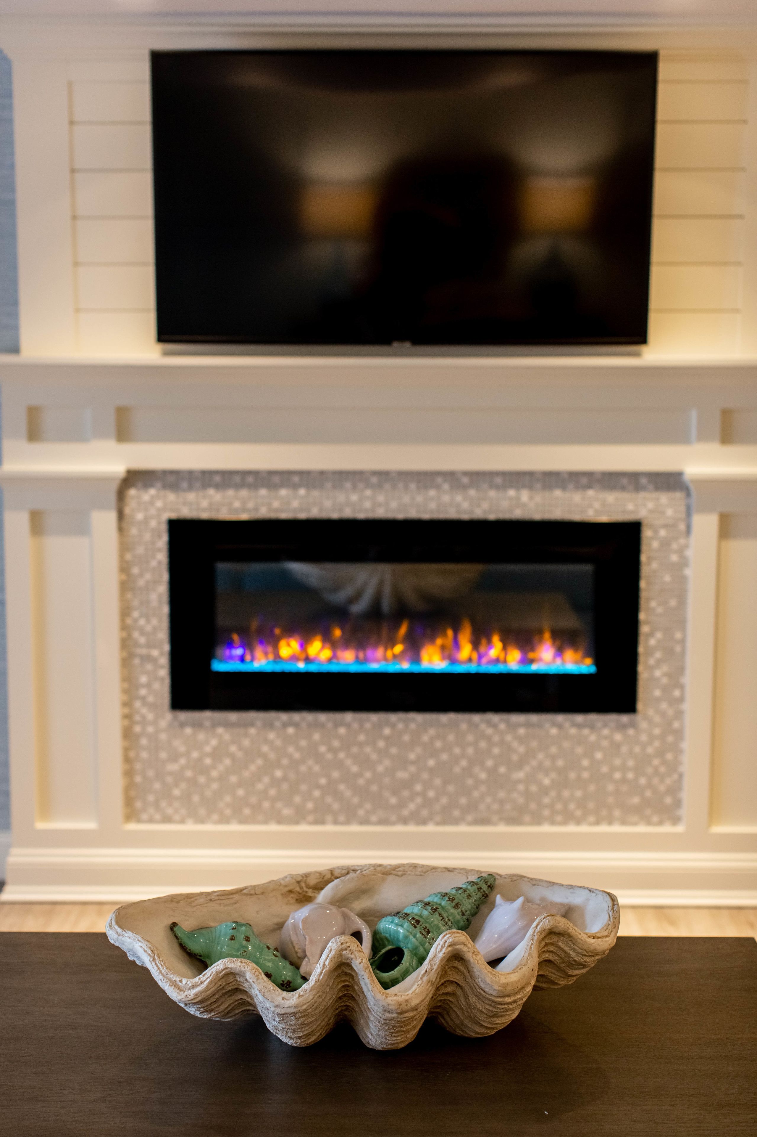 Una chimenea con un televisor montado encima, un cuenco decorativo sobre una mesa al frente y un papel tapiz estampado en la pared de fondo.