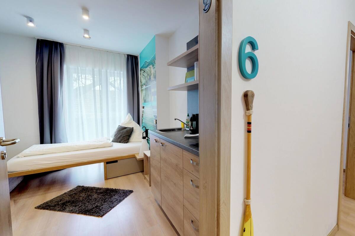 Una habitación de hotel moderna con cama, una kitchenette pequeña, un baño y un pasillo con un letrero de número.