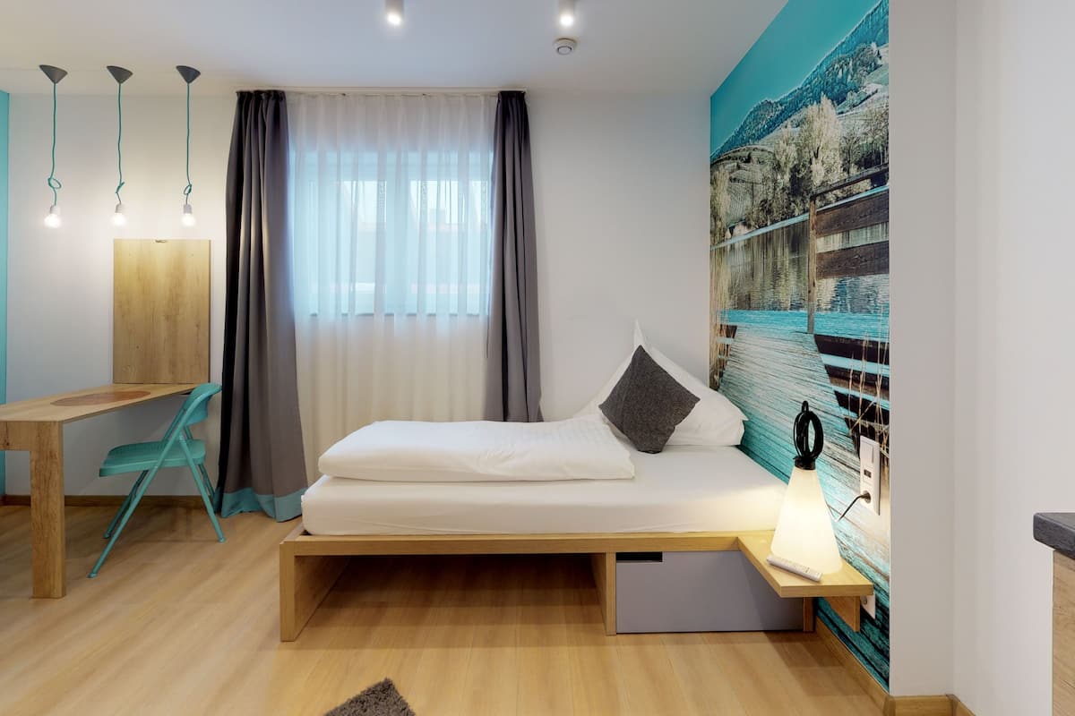 Un dormitorio moderno con cama, escritorio y silla. Hay un ventanal con cortinas, un televisor montado en la pared y un cuadro de un paisaje.
