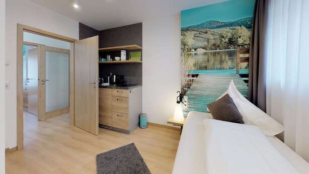 A modern hotel room with a bed, a small kitchenette, a painting of a lakeside scene, and a wooden door.