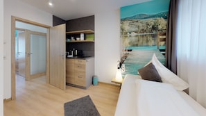 A modern hotel room with a bed, a small kitchenette, a painting of a lakeside scene, and a wooden door. - Pension Breitenauer See (Loewenstein)