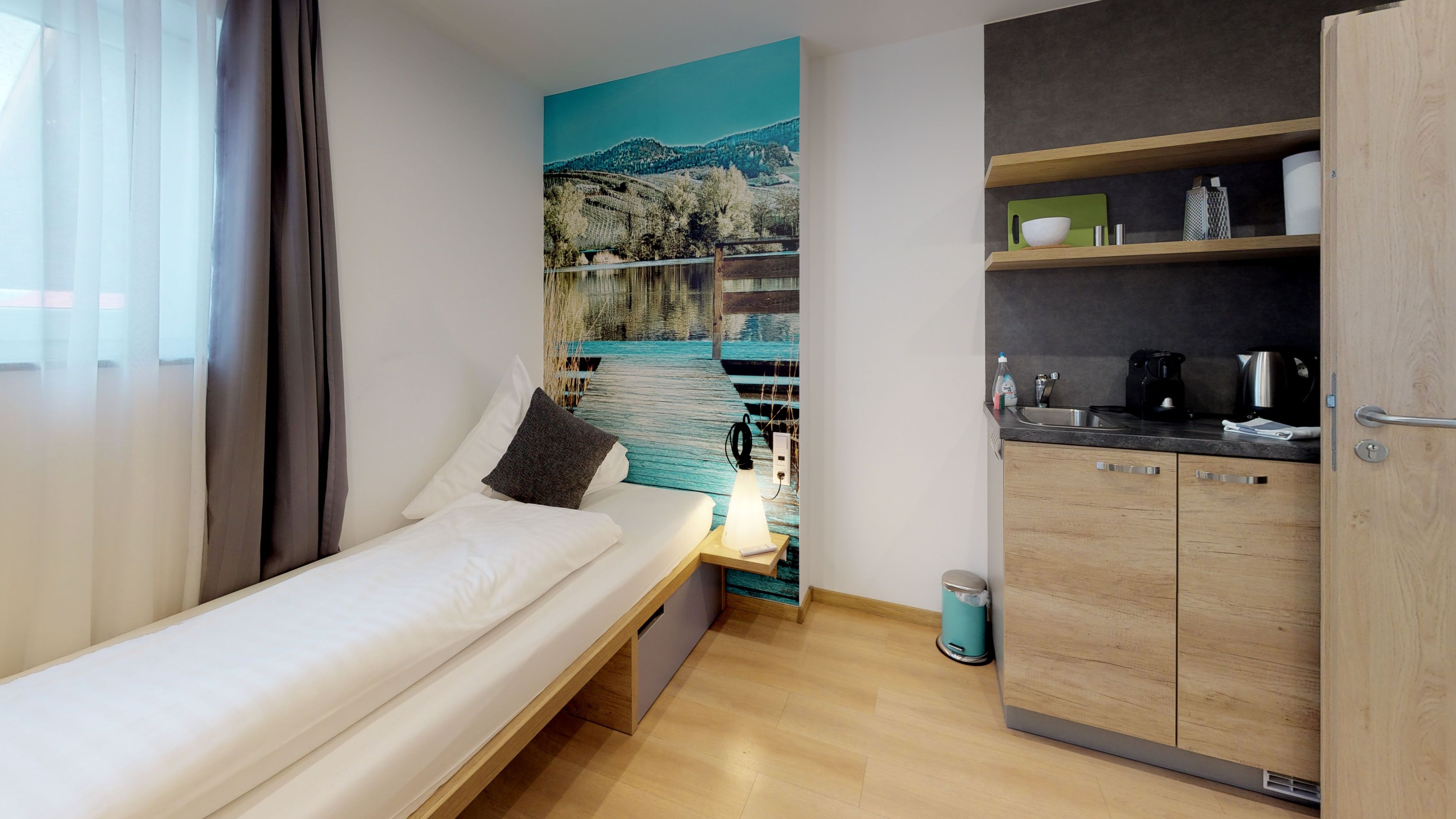 A compact room with a bed, a small kitchenette, and a large window with a scenic view.
