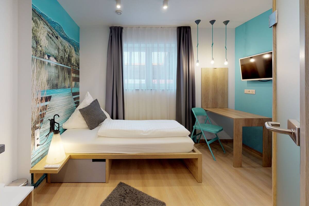 Una habitación de hotel moderna con cama, escritorio, silla, televisor y un ventanal con cortinas.