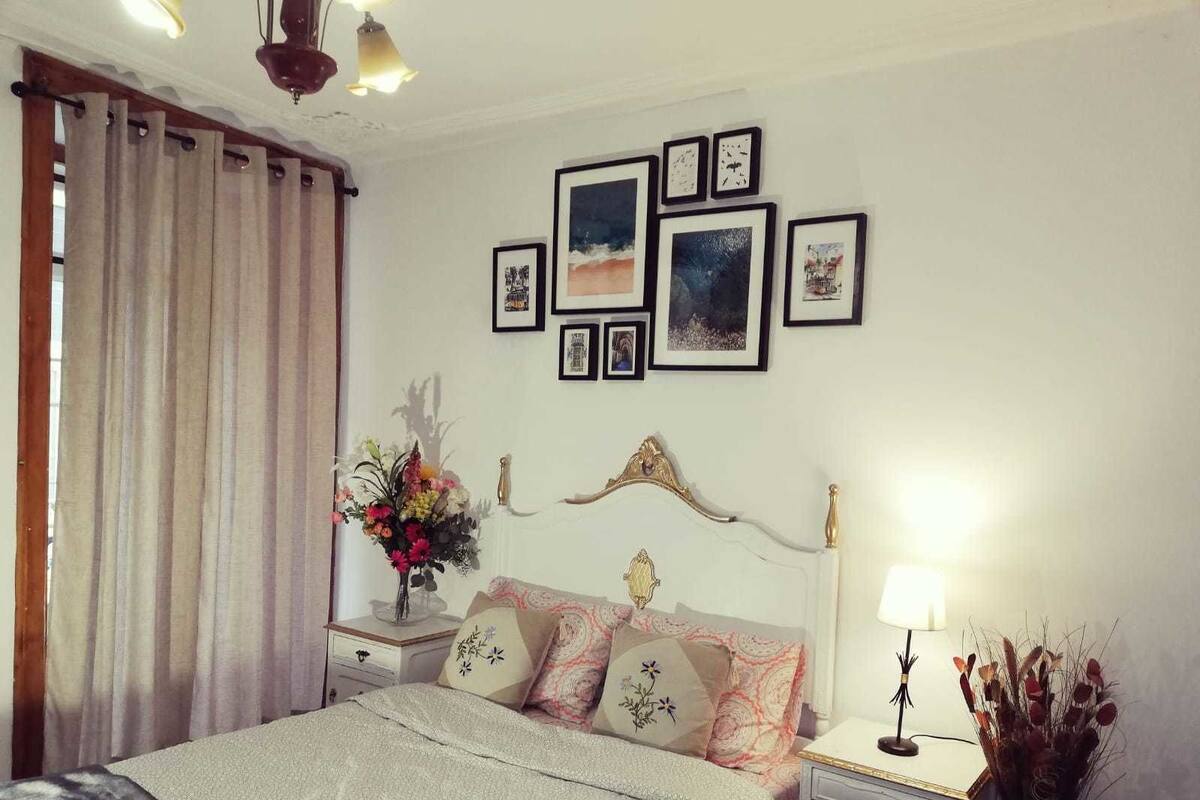 A bedroom with a bed, nightstands, a lamp, a vase of flowers, and framed pictures on the wall.