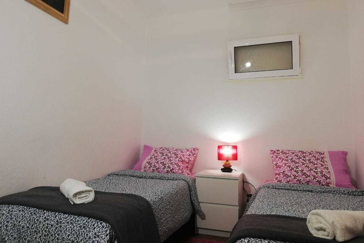 A small, simple room with two beds, a nightstand, a red lamp, and a window.