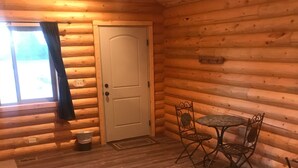 Una habitación tipo cabaña de madera con una puerta, una ventana con cortinas, una pequeña mesa con sillas y piso alfombrado.