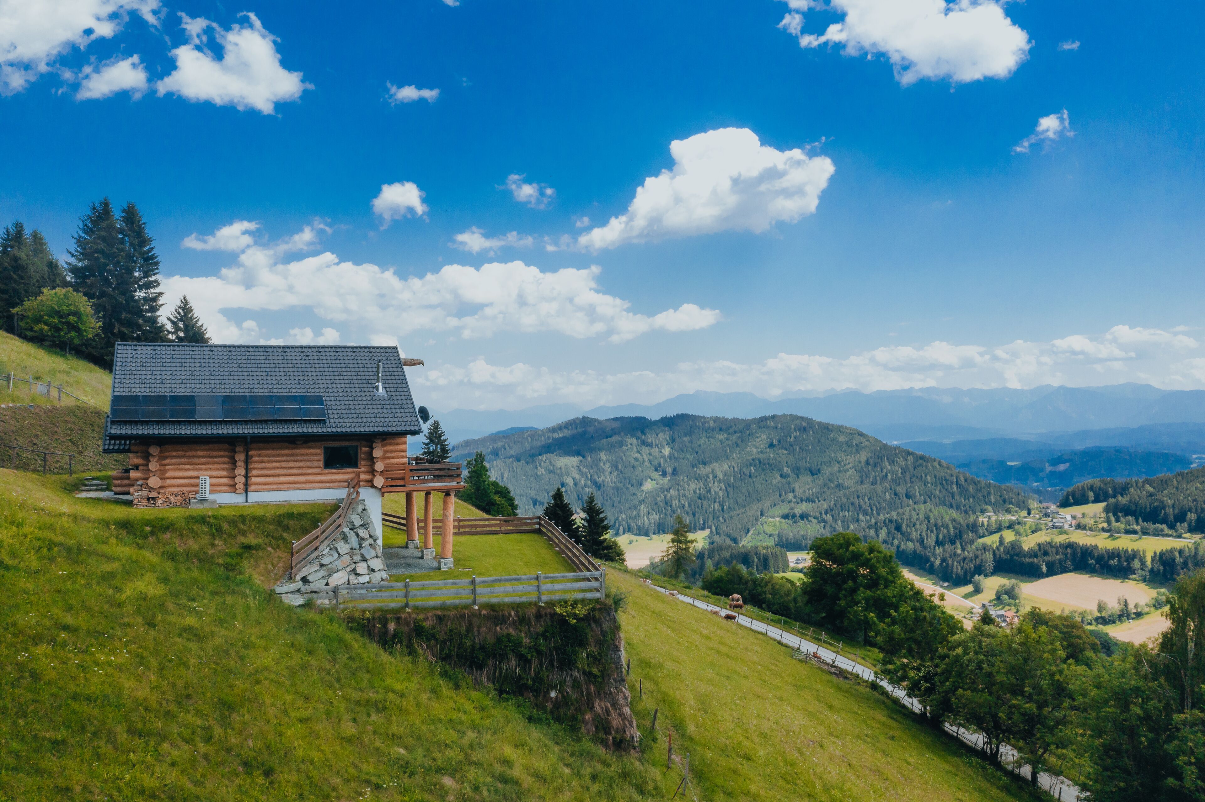A wooden cabin with a sloped roof, surrounded by a stone staircase and a wooden fence, overlooking a valley with mountains and a clear blue sky.