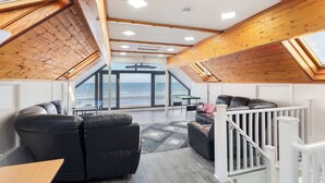 Un'ampia stanza con soffitto alto, travi in legno, una grande finestra con vista sul mare, un divano in pelle nera e una ringhiera bianca.