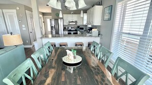 Un coin repas avec une table en bois, des chaises vertes, un lustre et une cuisine en arrière-plan.