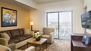Une chambre d’hôtel avec un canapé, un fauteuil, une petite table avec un vase, une lampe, une télévision et un balcon équipé de stores.