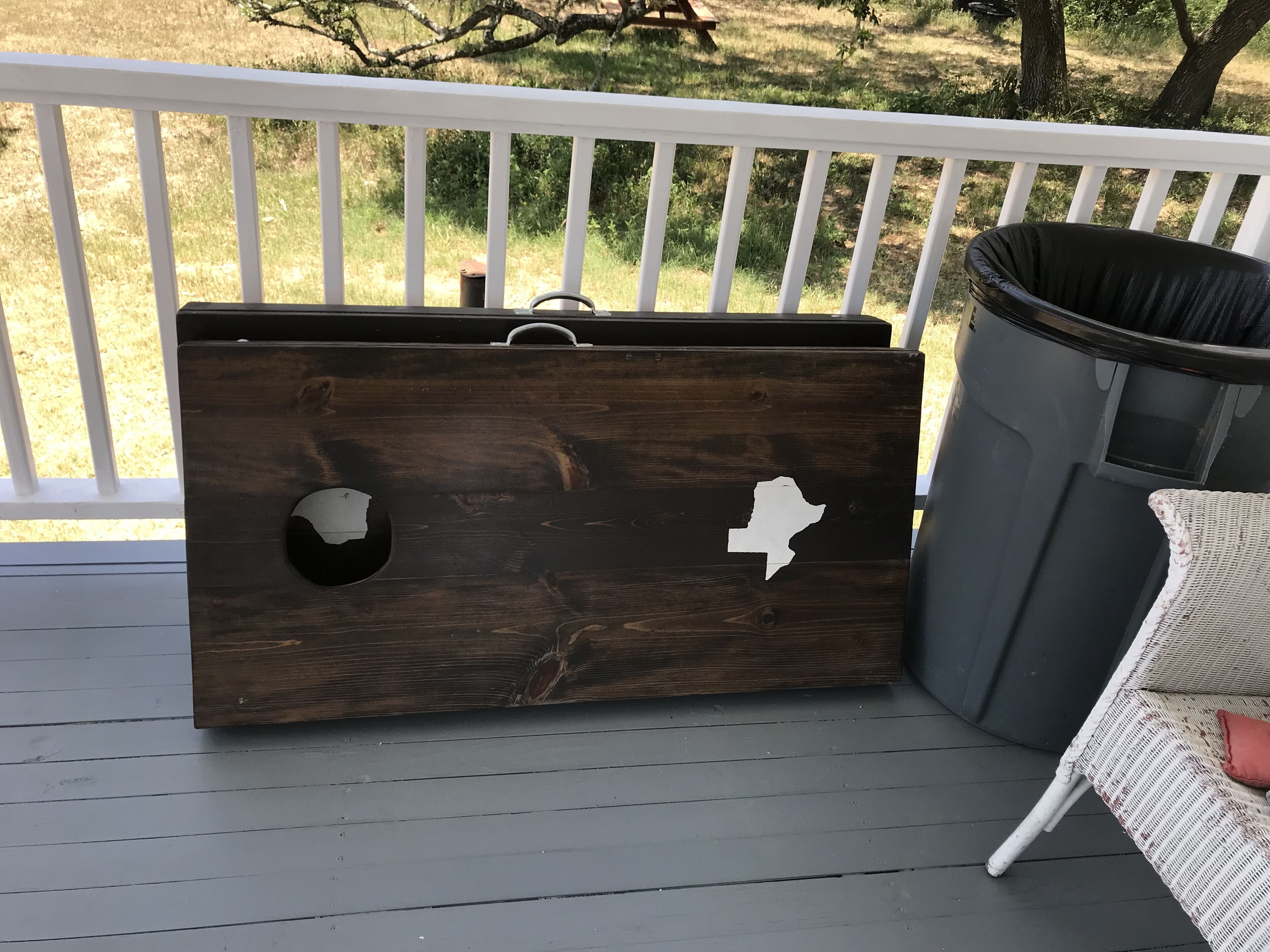 A wooden chest with a cut-out handle and a map design, placed on a deck with a wicker chair and a trash can nearby.