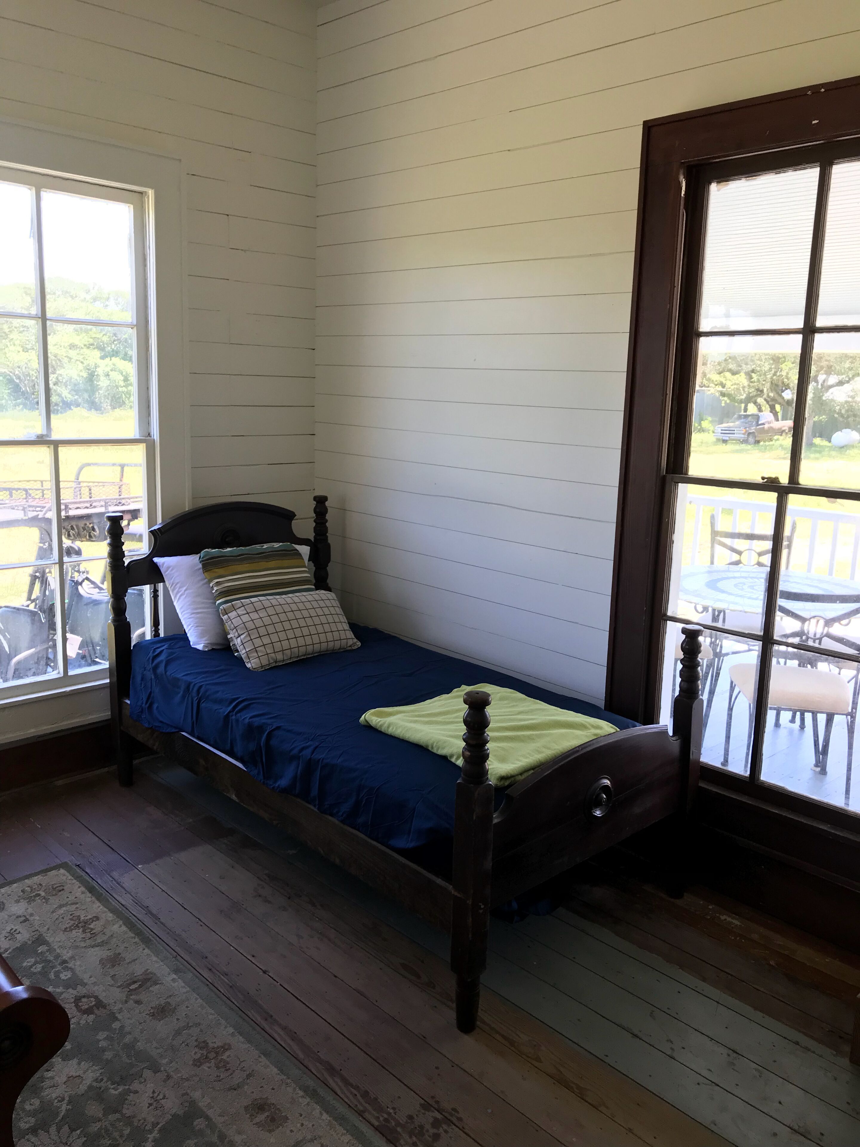 Una cama individual con estructura de madera y colcha azul en una habitación con paredes de paneles blancos y suelo de madera.