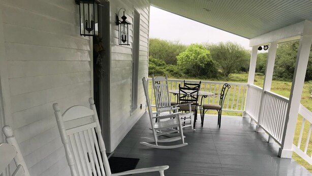 A porch with white rocking chairs and a table, white railings, and a view of greenery. - Koch Ranch - Hester House  (Columbus)