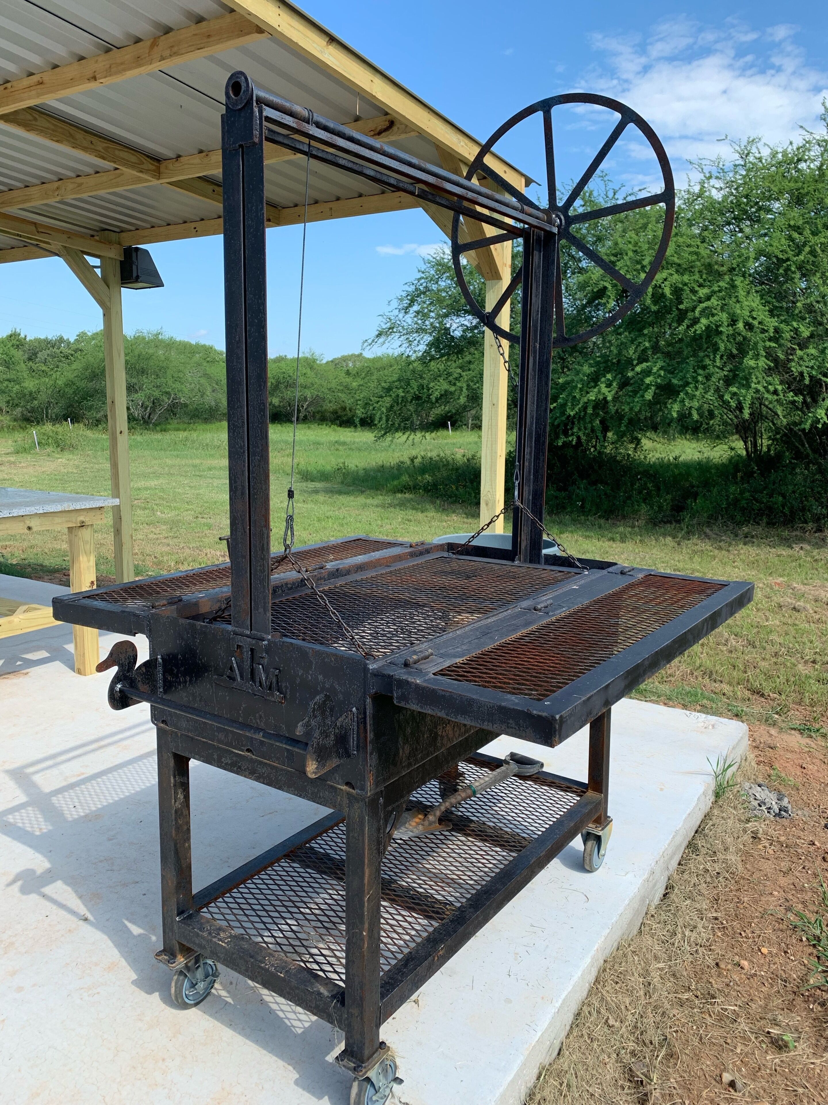 Un barbecue d’extérieur fixe, doté d’une grille en métal, de roues et d’un mécanisme de roues, installé sur une base en béton et placé sous un abri en bois.