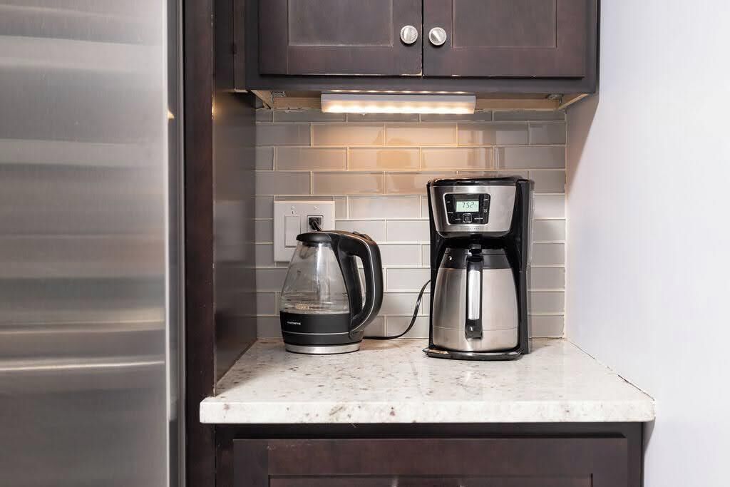 A kitchen with a coffee maker and a glass carafe on a marble countertop, under dark cabinets with a tiled backsplash.